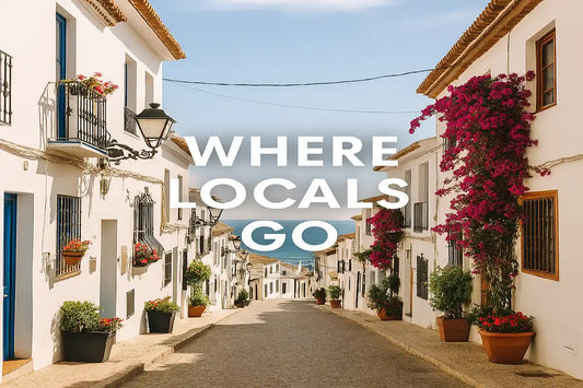 Sunlit Spanish village street with white buildings, potted flowers, and local pedestrians — a peaceful scene where locals live and relax away from crowds.